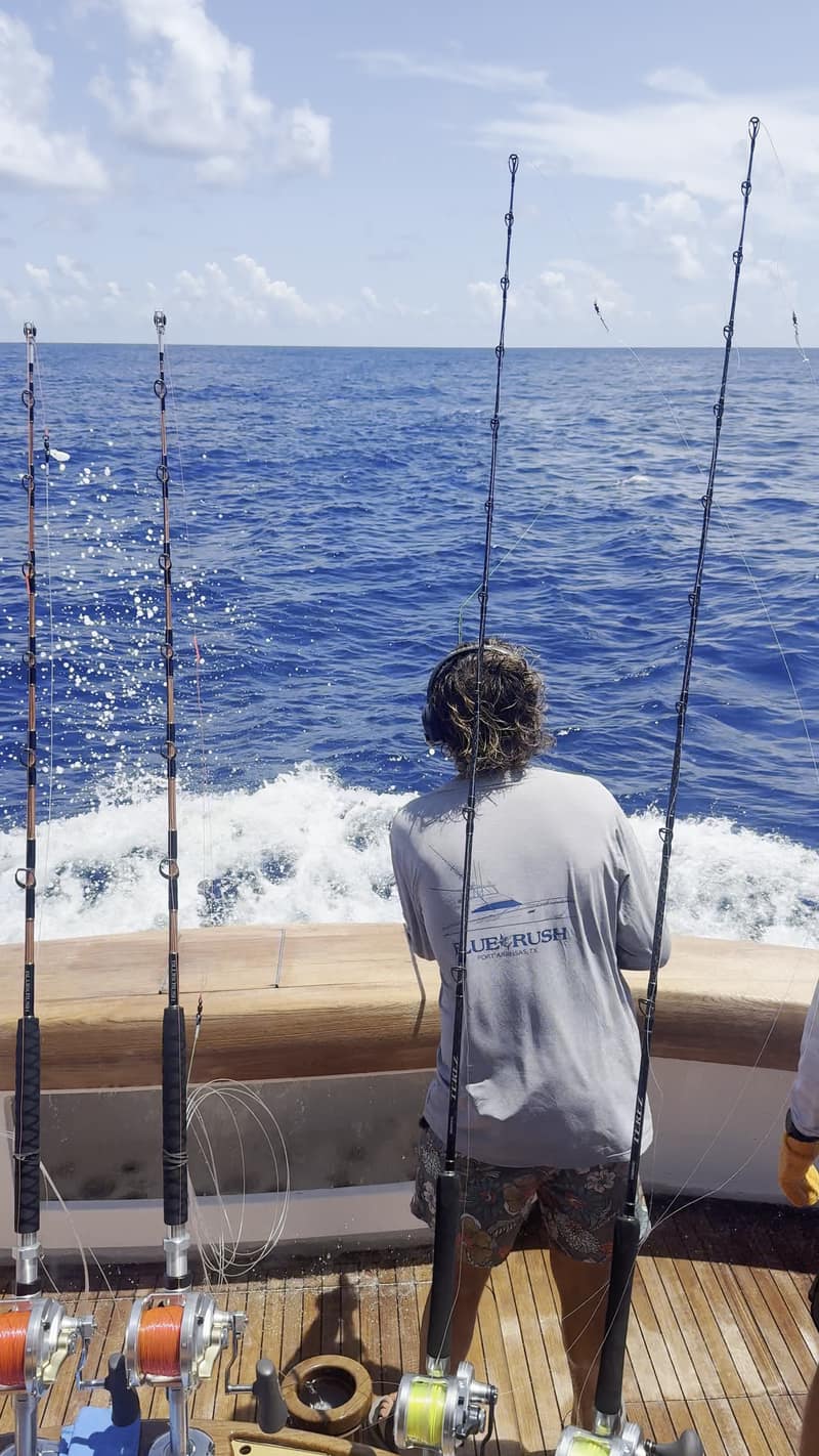 The cockpit of Blue Rush under way: rods standing in the covering board, reels spooled with red line, the white wake behind, and a crew member in a Blue Rush shirt watching the spread.