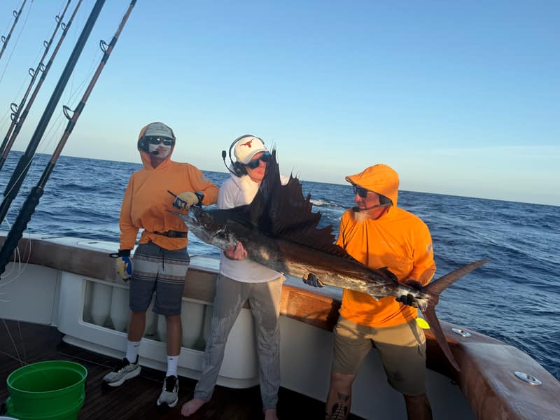 Three crew in orange shirts at the transom of Blue Rush holding a sailfish in golden evening light, rods racked behind them.
