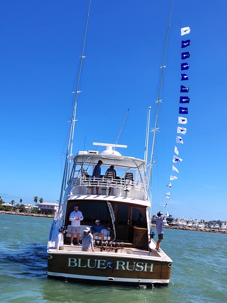 Blue Rush seen from astern coming into Port Aransas, a long string of blue and white release flags flying from the outrigger, the crew on the deck and the bridge.